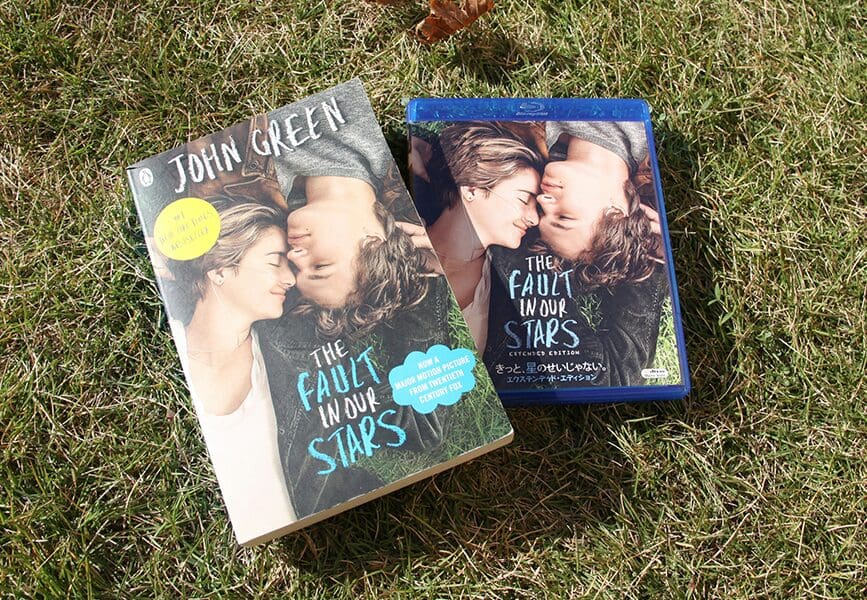A copy of the novel The Fault in Our Stars by John Green lies on grass next to the Blu-ray edition of its film adaptation. Both covers feature the same image of the two main characters lying close together, heads touching.