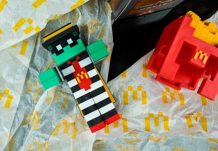 An example of a commercial tie-in, a McDonald’s-themed toy figure styled like a pixelated Minecraft character, wearing black-and-white stripes with a red tie featuring the McDonald’s “M” logo. The figure lies on McDonald’s-branded paper next to a red toy shaped like French fries.